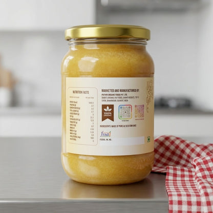 A glass jar of golden yellow ghee with a gold metal lid and a cream-colored label, set on a gray countertop with a red and white checkered cloth in the background.
